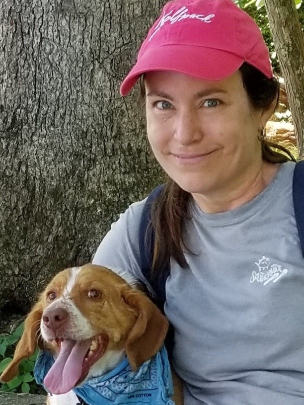 A photo of Brandye Smith-Goettler wearing a pink hat next to a tree and a holding a small cute panting dog